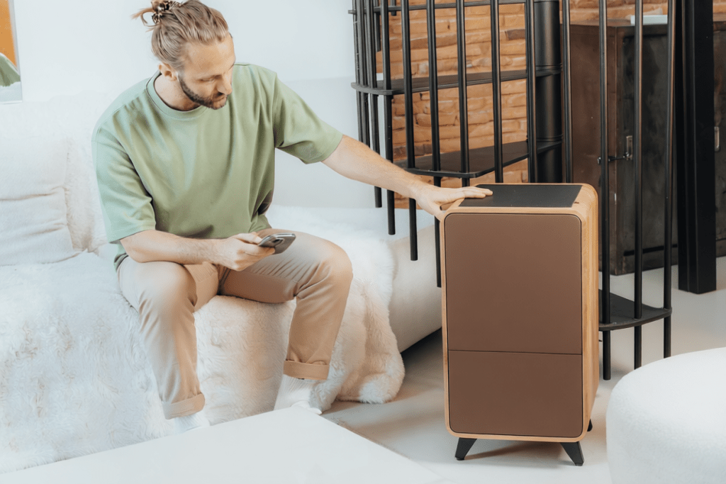 Un homme assis sur un canapé clair utilise un smartphone tout en se penchant vers un lombricomposteur moderne placé à côté de lui.